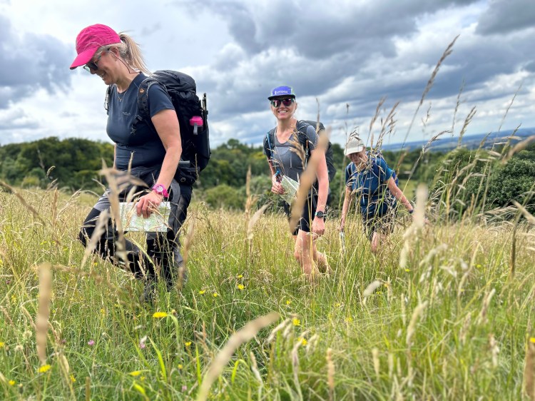three smiling ladies holding maps in their hands walking through a field of grass blowing in the gentle breeze, away from paths in the Chiltern Hills Oxfordshire and Buckinghamshire